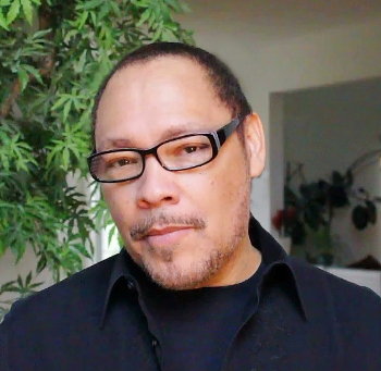 Photograph of author-Illustrator Floyd Cooper. He is  a light-skinned African American man, photographed from the shoulders up. He is wearing black glasses, a black collared shirt, and a black t-shirt underneath. 