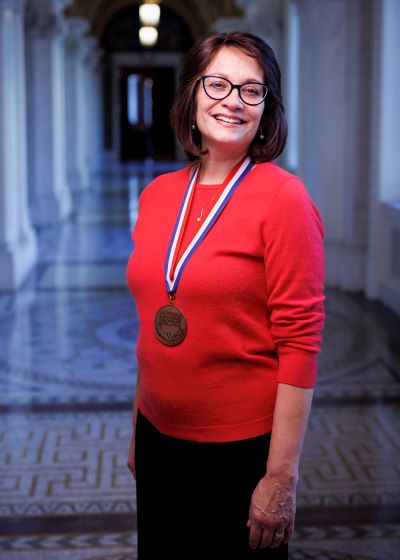 A vertical shot of author Meg Medina, standing and wearing the National  Ambassador for Young People's Literature Medal. She wears glasses and has shoulder length straight brown hair. She is wearing a red sweater and black pants. 