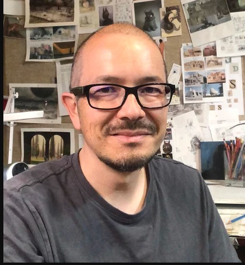 Author Shaun Tan. He is a thin biracial Asian man,  bald, and wears dark rimmed glassed. He  is wearing a faded black t-shirt. He is photographed from chest up in a seated position, and behind him is a bulletin board with several drawings tacked upon it. 