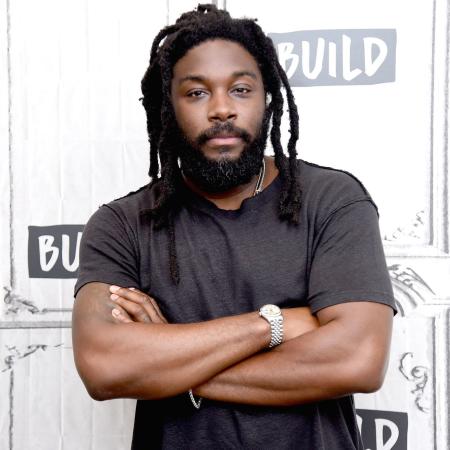 Photo shows a handsome black man with a beard and locked hair. He is wearing a short sleeve black t-shirt and has his arms crossed while looking directly at the camera. He stands againts a predominantly white background with some gray letter text as design elements. 